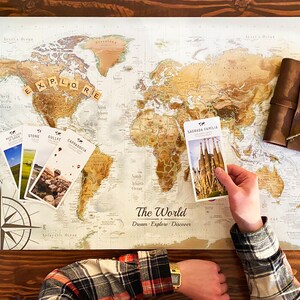 Puede incluir: Una persona est&aacute; usando un mapa del mundo para raspar y rastrear sus viajes. El mapa est&aacute; sobre una mesa de madera con una br&uacute;jula, un diario de cuero y varias postales. El mapa tiene las palabras "The World" y "Dream Explore Discover" impresas en &eacute;l. La persona est&aacute; sosteniendo una postal que dice "Sagrada Familia Barcelona Spain".