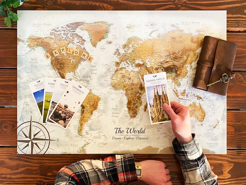 Puede incluir: Una persona est&aacute; usando un mapa del mundo para raspar y rastrear sus viajes. El mapa est&aacute; sobre una mesa de madera con una br&uacute;jula, un diario de cuero y varias postales. El mapa tiene las palabras "The World" y "Dream Explore Discover" impresas en &eacute;l. La persona est&aacute; sosteniendo una postal que dice "Sagrada Familia Barcelona Spain".
