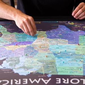 May include: A colorful map of North America with push pins marking different locations. The map is titled "Explore America" and is on a dark gray surface.