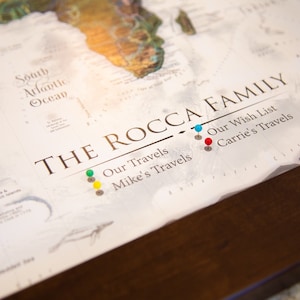 May include: A white world map with the text "THE ROCCA FAMILY" printed on it. The map has push pins in various colors, including green, yellow, blue, and red. The text "Our Travels", "Mike's Travels", "Our Wish List", and "Carrie's Travels" are printed below the family name.