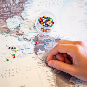 May include: A world map with push pins marking locations. The map is beige with brown outlines of countries. A hand is placing a yellow push pin on South America. A small clear container filled with colorful push pins is in the upper right corner of the image.