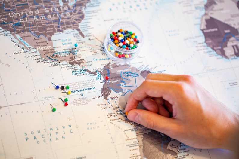 May include: A world map with push pins marking locations. The map is beige with brown outlines of countries. A hand is placing a yellow push pin on South America. A small clear container filled with colorful push pins is in the upper right corner of the image.