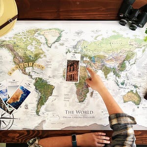 May include: A world map with the words "ADVENTURE" spelled out in wooden letter tiles. The map is surrounded by travel-themed items, including a safari hat, binoculars, and a leather-bound journal. The map has the words "THE WORLD" and "DREAM - EXPLORE - DISCOVER".