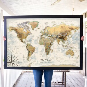 Puede incluir: Una persona sostiene un mapa del mundo enmarcado con un esquema de color dorado y marr&oacute;n. El mapa est&aacute; titulado "El Mundo: Sue&ntilde;a, Explora, Descubre".
