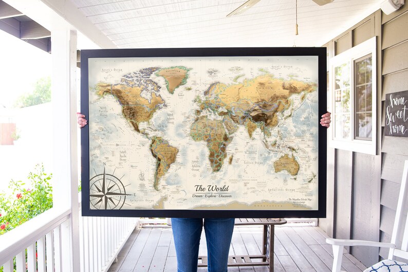 Puede incluir: Una persona sostiene un mapa del mundo enmarcado con un esquema de color dorado y marr&oacute;n. El mapa est&aacute; titulado "El Mundo: Sue&ntilde;a, Explora, Descubre".