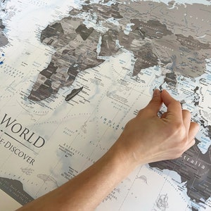 May include: A gray and white world map with blue oceans and continents. The map is titled "The World Dream Explore Discover". A hand is pointing to a location on the map. There are blue push pins on the map.