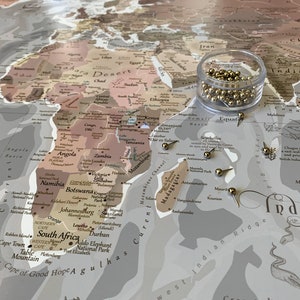 May include: A close-up of a world map with gold push pins marking locations. The map is printed on a light brown background with a beige and brown color scheme. The map shows the continents of Africa, Asia, and Australia. The map also includes the names of countries and cities.