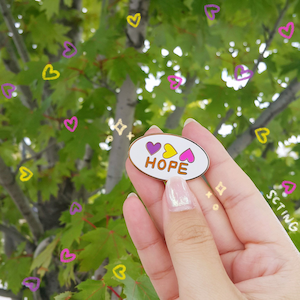 May include: White oval enamel pin with the word "HOPE" in gold lettering and three hearts in purple, yellow, and pink. The pin is being held by a hand against a background of green leaves and hearts.