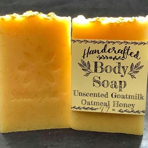May include: Two rectangular bars of handcrafted body soap. The soap is a light yellow color with a textured surface and a floral design. One bar has a label that reads "Unscented Goatmilk Oatmeal Honey" and weighs 4.7 oz.