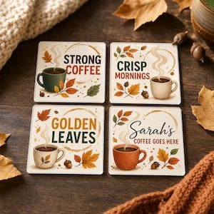 May include: A set of four square coasters featuring autumn-themed illustrations of coffee mugs, falling leaves, and acorns. Each coaster displays text: STRONG COFFEE, CRISP MORNINGS, GOLDEN LEAVES, and Sarah's COFFEE GOES HERE, with steam rising from mugs.