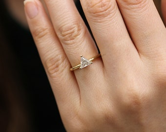May include: A close-up of a gold ring with a triangular, clear gemstone. The ring is on a finger, and the band is thin. The gemstone is set in a simple, elegant design. The ring is a piece of jewelry.