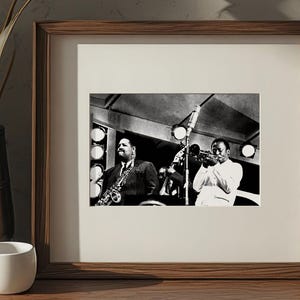 May include: A framed black and white photograph of two jazz musicians performing on stage. One plays a saxophone, the other a trumpet. The frame is a dark wood, and the photograph is displayed on a wooden surface.