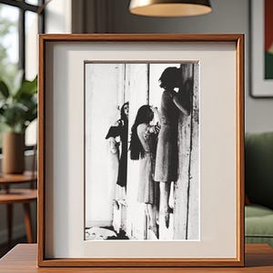 May include: A framed black and white photograph featuring three figures in dresses. The image is set against a white background within a wooden frame. The photograph is displayed on a wooden table, with a window and plant visible in the background.