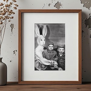 May include: Black and white framed photograph featuring a person in a bunny costume with two children. The photograph is in a wooden frame with a white mat. A vase with dried flowers is on a wooden surface.