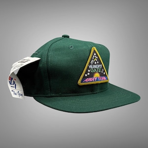 May include: A dark green baseball cap with a flat brim. The cap features a triangular patch with the text "Heaven's Gate Away Team" and a sun graphic. A white tag is attached to the side.