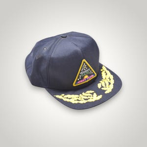 May include: A dark navy blue baseball cap featuring a triangular patch on the front with the text HEAVEN'S GATE AWAY TEAM and a sunrise graphic. The flat brim is decorated with yellow embroidered oak leaf clusters on both sides of the center.