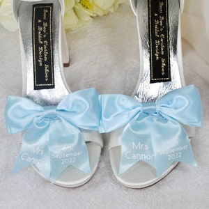 Puede incluir: Un par de zapatos de novia de satén blanco con lazos azul claro y cintas personalizadas que dicen "Mrs. Cannon" y "10 de septiembre de 2022". Los zapatos están decorados con una etiqueta negra que dice "Becci Boo's Custom Shoes & Bridal Design".