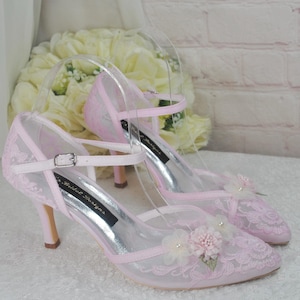 May include: A pair of pink lace bridal shoes with a delicate floral embellishment. The shoes have a pointed toe and a high heel.