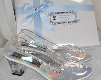 Könnte beinhalten: Ein Paar transparente, hochhackige Glasschuhe mit glitzerndem Design. Die Schuhe haben eine silberne, irisierende Oberfläche und sind mit Strasssteinen verziert. Die Schuhe befinden sich in einer Schachtel mit einem blauen Band und einem Etikett mit der Aufschrift "Disney Princess Inspired Collection".