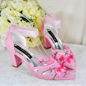 May include: Pink high-heeled shoes with a delicate floral embroidery design and a ribbon ankle strap. The shoes have a block heel and a pointed toe.