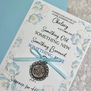 Puede incluir: Una tarjeta de boda con detalles florales azules y el texto "Felicidades en tu día de boda Chelsey 4 de octubre de 2025". Incluye un dije de moneda plateada con una cinta azul claro y las palabras "Something Old, New, Borrowed, Blue, and Sixpence for Her Shoe."