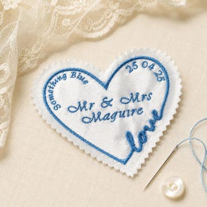 Puede incluir: Un parche de tela blanco en forma de corazón con bordes dentados y bordado azul. El texto dice Something Blue, 25 04 25, Mr & Mrs Maguire y love. Una aguja de coser con hilo azul y un botón blanco se encuentran cerca.