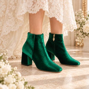 May include: A pair of emerald green velvet ankle boots with a block heel and a gold-toned zip pull on the inner side. The boots feature a smooth, rounded toe design and a soft, reflective fabric texture that catches the light.