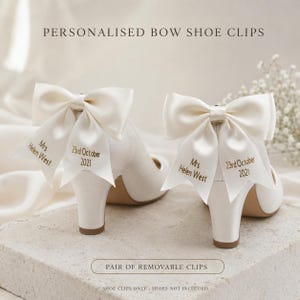 May include: A pair of ivory satin bow shoe clips featuring custom embroidery. Each ribbon tail displays the text Mrs Helen West and 23rd October 2021 in gold lettering. These removable accessories are designed to attach to footwear for decoration.