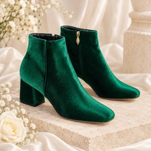 May include: A pair of ankle boots crafted from deep emerald green velvet material. These boots feature a block heel, a rounded toe, and a side zip closure accented with a gold-toned leaf-shaped zip pull for a secure and comfortable fit.
