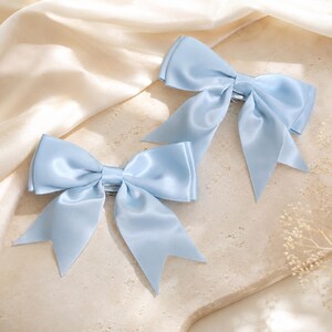 May include: Two light blue hair bows crafted from a smooth, satin-like fabric. Each bow features a central knot and two long, pointed tails extending downward. The bows are identical in color and construction, showcasing a soft, reflective sheen.