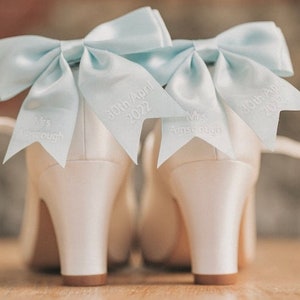 May include: A pair of white satin bridal shoes with light blue bows and personalized details. The bows are tied with a knot and the shoes are decorated with the text "Mrs. Amescough" and "30th April 2022".