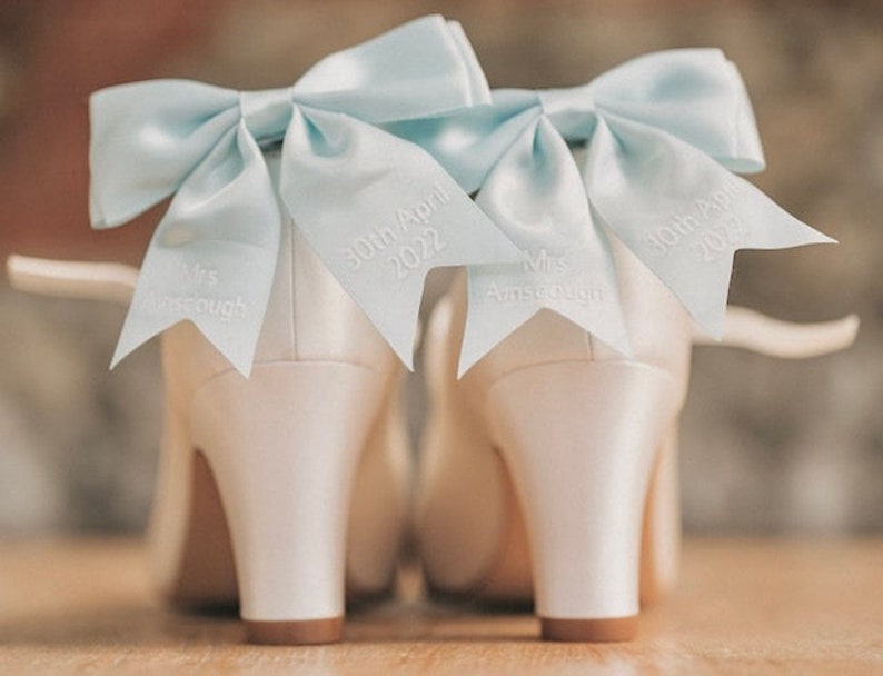 May include: A pair of white satin bridal shoes with light blue bows and personalized details. The bows are tied with a knot and the shoes are decorated with the text "Mrs. Amescough" and "30th April 2022".