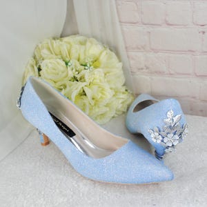 May include: A pair of light blue glitter heels with silver floral embellishments. The heels have a pointed toe and a low block heel.