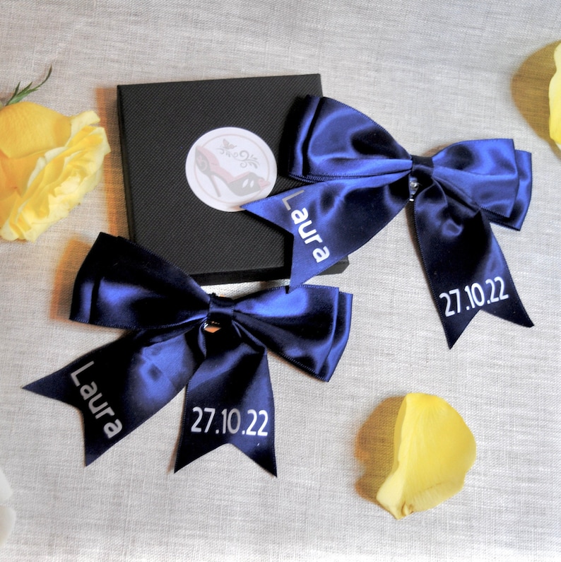 May include: Two navy blue satin bows with the name "Laura" and the date "27.10.22" embroidered in white on each bow.