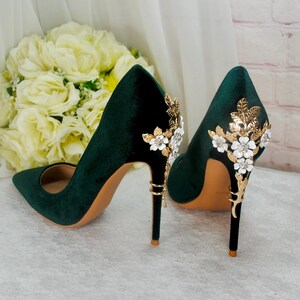 May include: A pair of dark green velvet high heel pumps with gold and white floral embellishments on the heel.