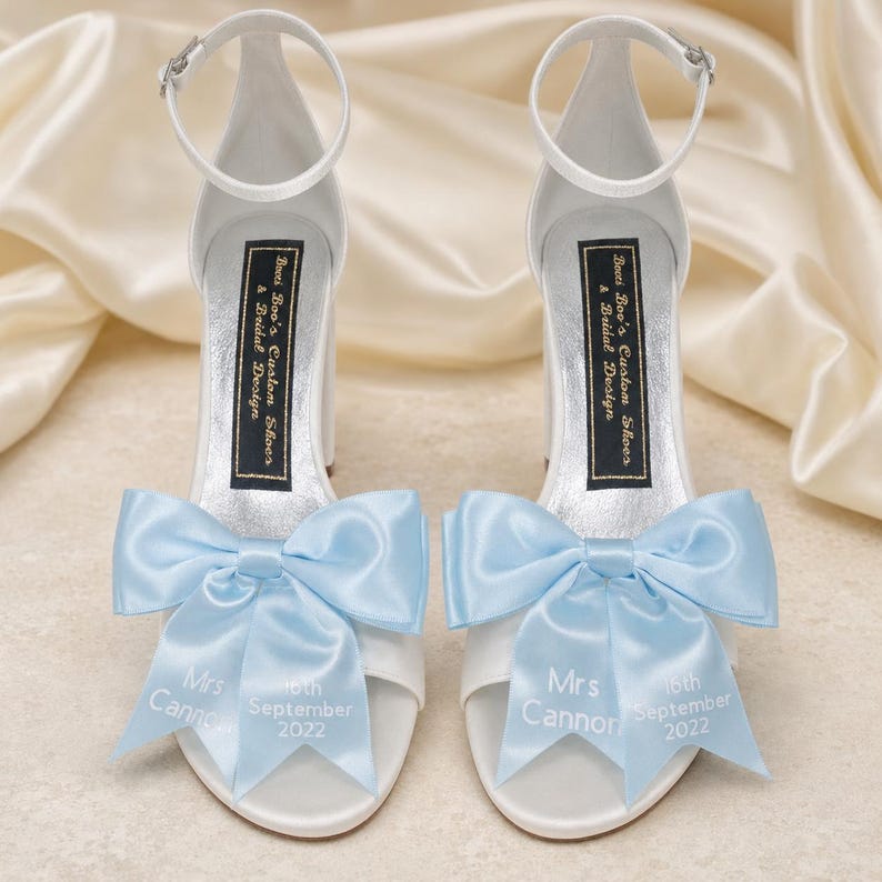 May include: A pair of white high-heeled sandals with ankle straps and large light blue satin bows on the toes. The bows feature the white printed text Mrs Cannon and 16th September 2022. The inner soles have a black label reading Boo's Custom Shoes & Bridal Design.