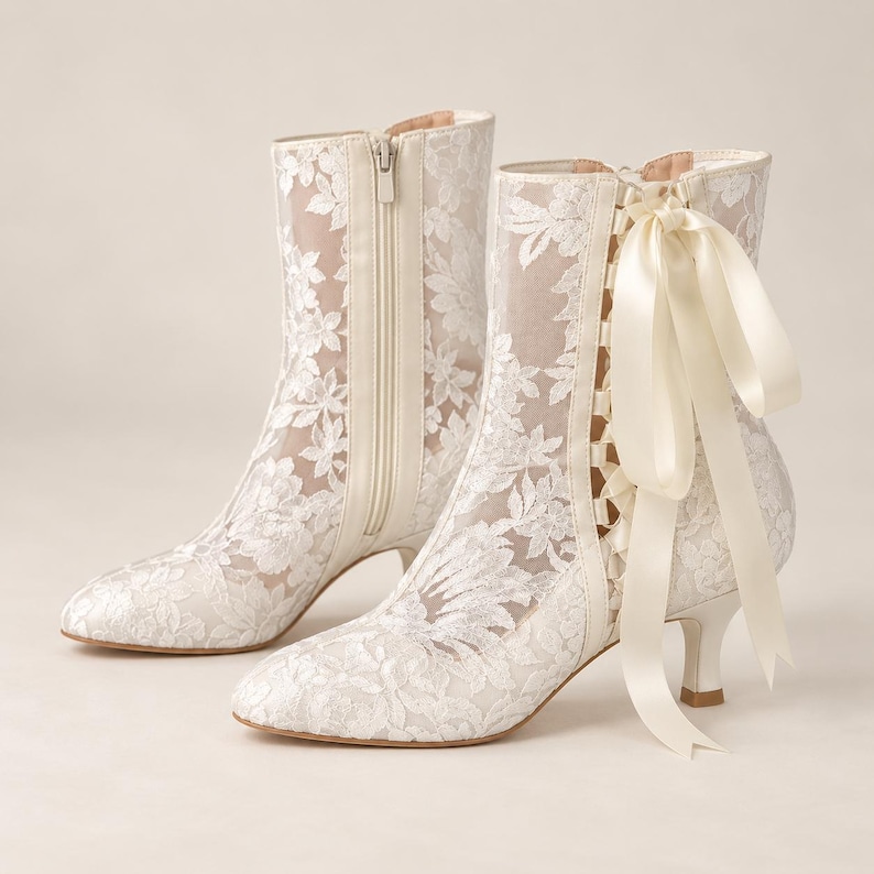 May include: A pair of white ankle boots featuring a sheer mesh base with intricate white floral lace overlays. The boots have a pointed toe, a low kitten heel, a side zipper closure, and a decorative satin ribbon lace-up detail on the side.