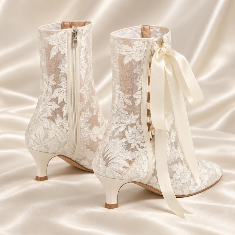 May include: A pair of ivory ankle boots featuring a sheer mesh overlay with white floral embroidery. The boots have a low kitten heel, a side zipper closure, and a decorative satin ribbon lace-up detail on the side with a tied bow at the top.