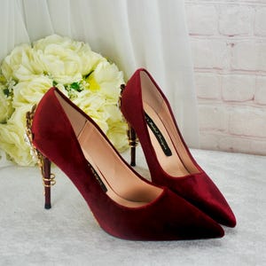 May include: A pair of burgundy velvet high heels with gold embellishments on the heel. The heels are pointed toe and have a stiletto heel.