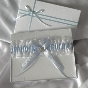 May include: A light blue garter with white lace trim and a rhinestone embellishment. The garter is personalized with the text "Danielle & Ashley" and "6th March 2025".