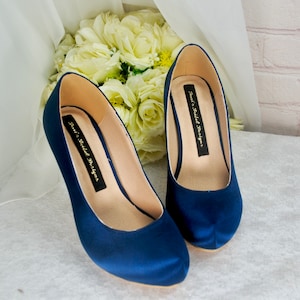 Op de afbeelding: Een paar donkerblauwe satijnen hoge hakken met een spitse neus. De hakken zijn gelabeld "Teal's Bridal Designs".