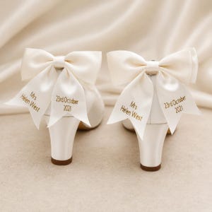 May include: A pair of ivory satin heels featuring large decorative bows on the back. The ribbons are embroidered with the text Mrs Helen West and 23rd October 2021 in gold thread. The shoes have a block heel and a smooth, polished finish.