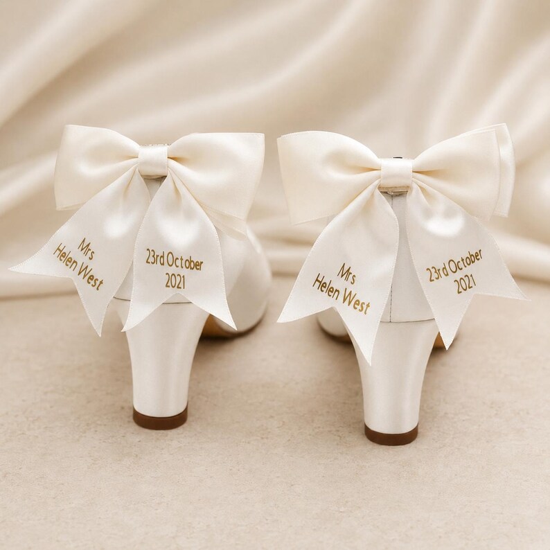 May include: A pair of ivory satin heels featuring large decorative bows on the back. The ribbons are embroidered with the text Mrs Helen West and 23rd October 2021 in gold thread. The shoes have a block heel and a smooth, polished finish.