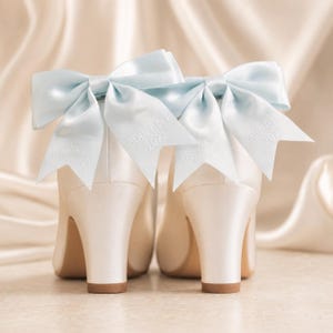 May include: A pair of ivory satin pumps featuring light blue ribbon bows attached to the heels. The ribbons are embroidered with the text Mrs Ainscough and 30th April 2022 in white thread. The shoes have a block heel and a smooth satin finish.