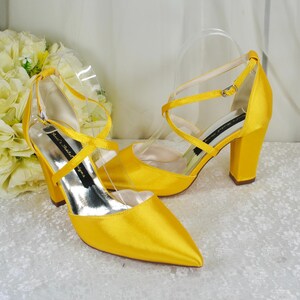 Puede incluir: Un par de zapatos de tacón de satén amarillo brillante con punta afilada, tacón de bloque y tiras finas cruzadas en el tobillo con una pequeña hebilla metálica. La suela interior muestra el texto Ricci's Bridal Designs en letra cursiva.