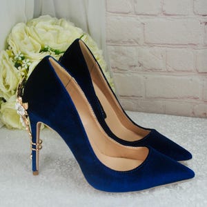 May include: A pair of blue velvet high heels with gold embellishments on the heel. The heels have a pointed toe and a stiletto heel.