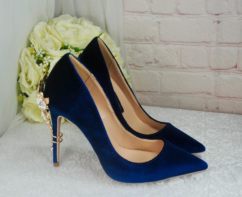 May include: A pair of blue velvet high heels with gold embellishments on the heel. The heels have a pointed toe and a stiletto heel.