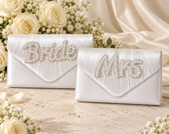 Pode incluir: Duas bolsas de mão tipo envelope em cetim branco com detalhes em tecido plissado. Uma exibe a palavra Bride em letras incrustadas com pérolas, enquanto a outra exibe a palavra Mrs em letras cursivas correspondentes com pérolas.