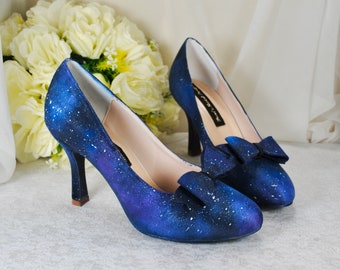 Könnte beinhalten: Ein Paar blaue High-Heel-Schuhe mit Galaxie-Druck und einer weißen Schleife an der Spitze.