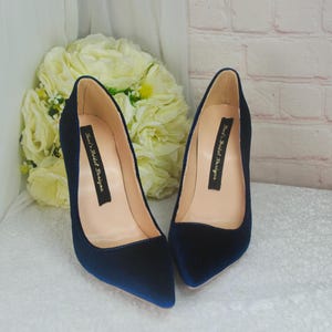 May include: A pair of navy blue velvet pointed-toe pumps with a stiletto heel. The shoes have a label on the inside that reads "Rex's Bridal Designs".